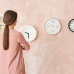Una donna con i capelli lunghi di spalle, osserva quattro orologi sulla parete appesa che ha di fronte a lei. Gli orologi segnano quattro orari diversi.