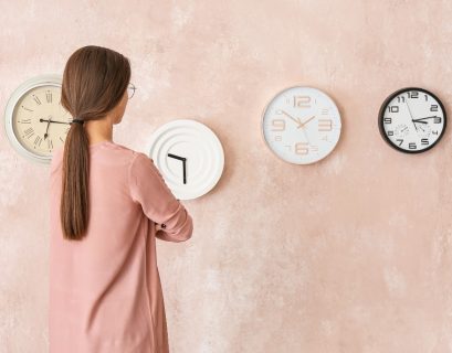 Una donna con i capelli lunghi di spalle, osserva quattro orologi sulla parete appesa che ha di fronte a lei. Gli orologi segnano quattro orari diversi.